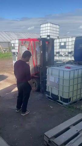A man in a maroon shirt and black pants stands to the left of a red forklift. The forklift is lifting a white cubic container on a pallet. The forklift has a clear enclosure over the driver's seat. In the background, there are more white cubic containers and greenhouses. The ground is dirt.