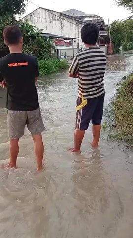Two men stand ankle-deep in floodwater on a street in a residential area. One man wears a black shirt and gray shorts, and the other wears a striped shirt and blue shorts. The camera pans to show more of the flooded street, where two young women are walking through the water. The water appears murky and obscures the road surface. Buildings and trees line the street, and the sky is overcast.
