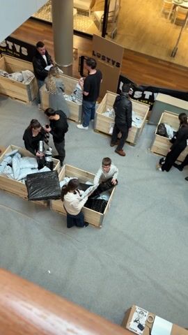 A high-angle view depicts a group of people gathered around several wooden crates filled with items in black plastic bags and white packages. Most of the people are adults, but there is a young boy wearing glasses who is also digging through a crate. The adults are interacting with each other while examining the contents of the crates. The crates are labeled with the logo "LOOTYS." The setting appears to be an indoor space, possibly a mall or event area, with a wooden floor visible in the background. People are walking in the background.