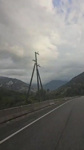 The video shows a view from a moving vehicle on a road. The road curves to the right, and a concrete barrier runs along the left side. Beyond the barrier is a landscape with rolling hills and mountains covered in green trees. In the distance, the mountains are shrouded in clouds, and the sky is overcast with a mix of white and dark clouds. A wooden utility pole with wires stretches across the upper portion of the frame, with several wires extending out from it.