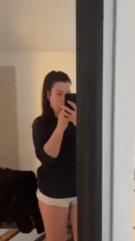 A woman stands in front of a mirror brushing her teeth while holding a smartphone. She is wearing a dark-colored, long-sleeved top and light-colored shorts. Her dark hair is pulled back in a ponytail. The mirror has a black frame. A rack of clothes is visible in the background to the left. The room appears to be a bedroom or hotel room.