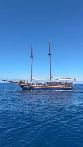 A large, two-masted schooner-style boat named "BONITA DA MADEIRA" is shown sailing on clear blue ocean waters under a brilliant, cloudless sky. The boat is blue with wooden accents and features two tall masts with sails rolled up. Many people are visible standing on the boat's deck, observing the surroundings, particularly towards the bow. The vessel is actively moving, leaving a white wake behind it. A Portuguese flag can be seen flying from the stern. The scene depicts a bright, sunny day at sea, likely off the coast near Caniço, Portugal, with passengers enjoying a calm cruise.