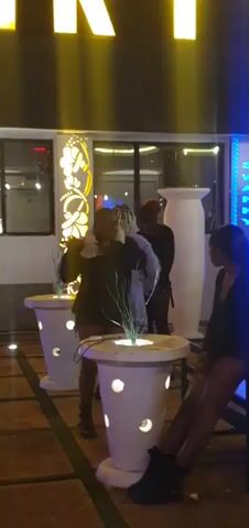 A group of people are gathered around decorative planters on a patio at night. A woman in the foreground is wearing a dark jacket and a white fur collar, and is gesturing with her left hand. Two other people are standing behind her, and another person is seated to the right. The area is lit with yellow and blue lights.