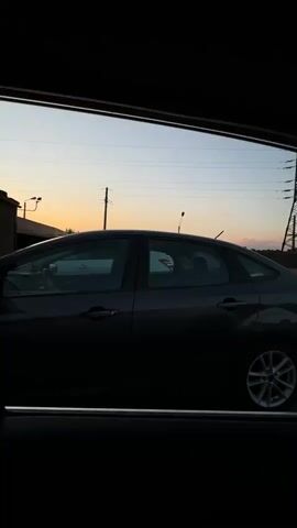 The video shows the interior of a car looking out through the windshield and side window at a parking lot. The time of day appears to be sunset or dusk. In the foreground, the steering wheel and dashboard of the car are visible. Through the windshield, several cars are parked in rows. A large electrical tower can be seen in the distance. The side window reveals another car parked alongside the vehicle from which the video is filmed. The sky is visible with some clouds and power lines.