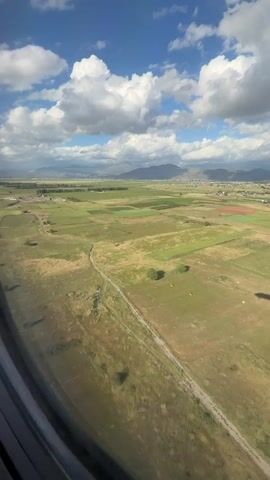 The video shows a view from an airplane window looking down at a rural landscape. The terrain consists of green and brown fields, some of which appear to be farmland with rows of crops. In the distance, there are mountains visible under a blue sky with scattered white clouds. The shadow of the airplane can be seen on the ground. The camera is relatively stable, offering a clear view of the scene below.
