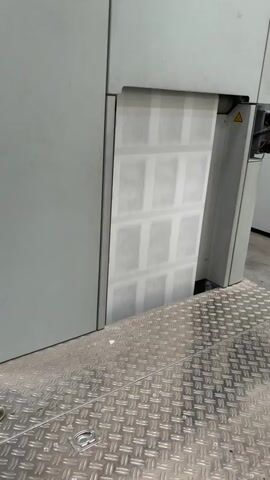 A large printing press is visible in the frame. A wide band of paper emerges from the machine, imprinted with a repeating grid pattern of rectangular shapes. The paper is predominantly white with light gray markings within the grid. The press is painted in shades of gray. A metal platform with a diamond plate texture is in the foreground. A yellow warning sign is affixed to the side of the press.