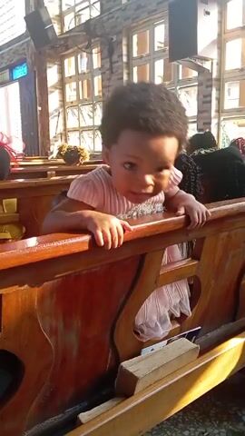 A little girl in a pink dress stands up in a church pew, holding onto the back. She looks around and smiles. The camera then pans around, revealing the rest of the row in the pew, which includes other children and adults. The church has a wall of windows behind the pews.