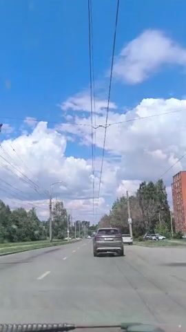 The video shows a view from a vehicle traveling on a road. The sky is blue with white clouds. There are overhead wires, likely for an electric vehicle or trolleybus. Several cars are visible on the road ahead and to the side. Trees line the right side of the road, and a building is visible in the distance on the right. There are streetlights and utility poles along the road. The camera is positioned at the front of the vehicle, suggesting a dashcam view.