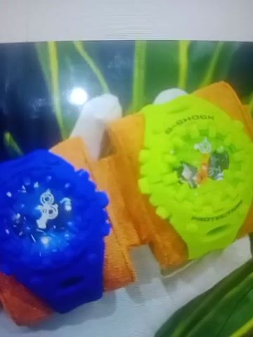 The video displays a close-up shot of two wristwatches, one blue and one neon green, placed on orange fabric. The watches appear to be G-Shock models, with the green watch clearly showing the "G-SHOCK PROTECTION" branding. The blue watch has a white design on its face that is difficult to discern due to lighting. The background consists of blurry green foliage. The lighting is bright, with a strong glare on the left side of the image. The video quality is low, with a blurry and pixelated appearance.