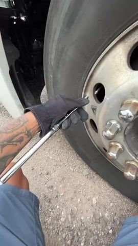 A person wearing a black glove and blue shorts is using a long metal tool to inspect the tire of a large vehicle. The tool has a measurement scale on it. The person's left arm, which is covered in tattoos, is visible. The tire is a large truck tire, and the background shows gravel. The action appears to be the person measuring the depth of the tire tread or checking the air pressure.
