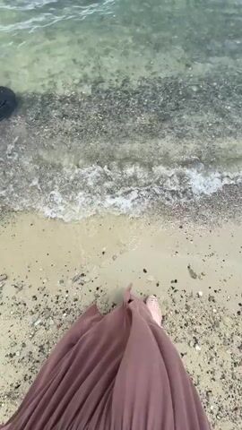 A person walks near the shore of a body of water. The water is clear and shallow, with small stones visible beneath the surface. The person is wearing a long, flowing brown skirt and light-colored shoes. Gentle waves lap at the shore, creating a foamy white line where the water meets the sand and small pebbles. The sky is a bright blue with a few scattered white clouds. In the distance, a single person can be seen swimming in the water. The scene is tranquil and appears to be a pleasant day at the beach or lake.