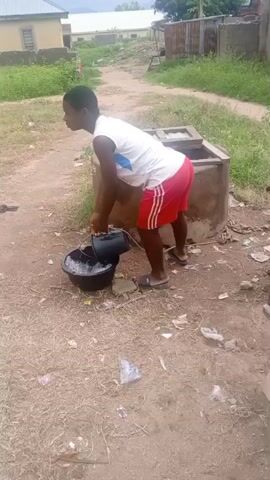 A person wearing a white tank top and red shorts with white stripes is standing next to a well. The person is pouring water from a black bucket into a larger black basin on the ground. The well has a concrete structure around it and a wooden cover. There is debris on the ground around the well, including plastic bags and paper. In the background, there are buildings and vegetation. The person appears to be in a rural or semi-rural environment.