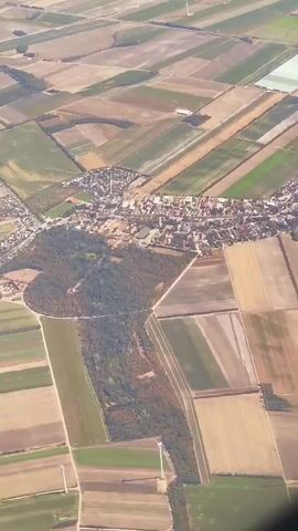 An aerial view captures a patchwork of agricultural fields and a small town. The fields are divided into various rectangular plots, some appearing to be recently harvested with brown soil, while others are green with crops. In the distance, several wind turbines stand tall against the horizon, their blades motionless. A cluster of buildings, forming a town or village, is nestled amidst the fields, with streets and houses visible from this elevated perspective. The scene suggests a rural landscape, likely in an area with active agricultural practices and renewable energy generation.