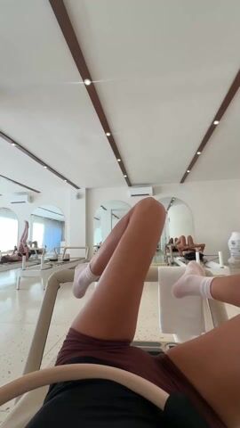 The video shows a person lying on a Pilates reformer, performing leg exercises. The camera is positioned from the person's point of view, looking upwards at the ceiling. Their legs are extended and moving in various positions, sometimes holding a Pilates ring. Other people are visible in the background, also using Pilates reformers in the same room, which is brightly lit with a white ceiling and light wooden beams. The walls are white with arched mirrors, creating an airy and spacious atmosphere. The person is wearing maroon shorts and white socks. A small tattoo is visible on one of their legs. A white towel is draped over the side of the reformer.