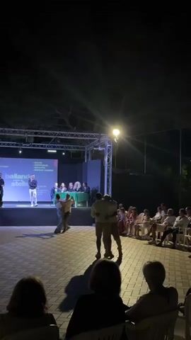 The video shows an outdoor evening event with a stage set up. On the stage, there's a screen displaying text and several people seated behind a table. In front of the stage, two couples are dancing. One couple is dancing center stage, and another couple is dancing slightly to the left. An audience is seated on chairs to the right of the stage, watching the performance. The overall ambiance suggests a dance competition or showcase.