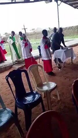 The video shows a group of children in church vestments standing in a line. Some children are wearing white robes with red sashes, while one child is wearing a green and white robe. They appear to be participating in a religious service. In the background, there is a table with a white cloth and some objects on it, possibly an altar. There are also adults present, one of whom is standing behind the table. The setting appears to be an outdoor or semi-outdoor space, with a corrugated metal roof overhead and sandy ground. Chairs are arranged in the foreground, some of which are occupied. The overall atmosphere suggests a religious ceremony or procession.