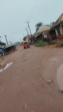 The video shows a muddy, unpaved road with puddles of water. Several houses and shops line the street, some appearing run-down. A colorful motorized tricycle is visible in the distance, driving away from the camera. Power lines stretch across the sky. The overall impression is of a somewhat neglected rural or semi-urban environment, possibly after rain.