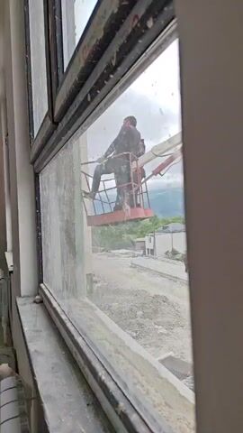 A person in a lifted basket is cleaning a window from the outside. The person is wearing camouflage clothing and appears to be using a tool to clean the window. The background shows a construction site with dirt and debris, and a mountainous landscape. The window is dirty and streaked. The overall scene suggests outdoor work on a building.