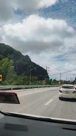 This is a first-person perspective video taken from inside a car driving on a road. The road appears to be a highway with at least two lanes in the direction of travel. There is a white car ahead in the right lane. On the left side of the road, there is a steep, tree-covered mountain. Utility poles with numerous wires are visible along the side of the road and extending into the distance. The sky is partly cloudy with large, white cumulus clouds against a blue sky. There are speed limit signs and directional signs visible along the road. The sun appears to be shining.