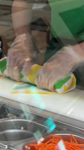 The video shows a close-up view inside what appears to be a Subway restaurant, focusing on a food service worker wrapping a sandwich. The worker is wearing a green uniform and clear plastic gloves. They are positioned behind a stainless steel food preparation counter that features several inset containers holding ingredients; clearly visible are chopped orange carrots and a green, possibly avocado-based, mixture. A white cutting board is on the counter directly in front of the worker.

The main action involves the worker swiftly and efficiently wrapping a long food item, presumed to be a sub sandwich, in a distinctive Subway-branded paper wrapper. The wrapper features a pattern of green, yellow, and white, with the word "SUBWAY" partially visible. The worker's gloved hands are seen tightly rolling and securing the paper around the sandwich. The video is shot through a transparent screen or glass barrier, causing some reflections and a slight diffusion of light in the foreground. The movement of the worker's hands is quick and practiced.