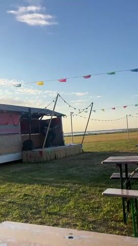 The video shows an outdoor setting with a small stage. A person is playing a guitar on the stage. The stage is decorated with what appears to be straw-like material and strings of lights. Behind the stage is a small structure that looks like a trailer or a shed. Colorful pennant flags are strung across the sky. In the foreground, there are picnic tables and chairs. The sky is blue with some clouds, and there is a body of water in the background. The lighting suggests it is either early morning or late afternoon.