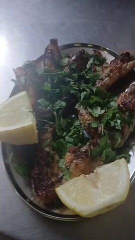 A top-down view shows a silver platter holding a meal on a metal table. The platter contains several pieces of grilled chicken, garnished with fresh green herbs, possibly parsley or cilantro. Two large wedges of lemon are placed on the platter, one on the left and one on the right, to the side of the chicken. The chicken pieces appear golden brown with some charred spots. The metal table reflects some light, indicating a polished or metallic surface.