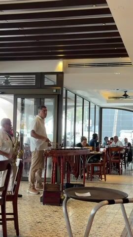 Inside a restaurant, two musicians are playing instruments for a seated audience. One musician, on the left, plays a saxophone, wearing a white shirt and light khaki pants. The second musician stands to his right, playing a marimba, wearing a white button-down shirt and khaki pants. The marimba has intricately carved red legs. People are seated at tables in the background, some eating, others talking. The restaurant has large windows and a decorative brown wooden ceiling. The floor appears to be tiled. A small round table with a white paper on it is in the foreground.