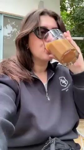 A young woman with long brown hair and sunglasses drinks from a glass of a tan-colored beverage. She is wearing a dark gray zip-up sweatshirt and sits outdoors, visible in the background are green plants and a building with a blue door. The camera focuses on the woman's face as she takes a sip, then she holds up the glass and speaks. The camera briefly moves off the subject, focusing on an unclear object.