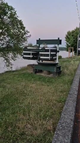 The video pans across a large industrial press-like structure situated on a grassy embankment next to a body of water, possibly a river. The structure, painted green and white, consists of two cylindrical barrels supported by a metal frame. Trees line the background on the opposite side of the water, and a path runs along the right edge of the frame. The sky is a muted twilight hue.
