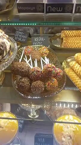 The video displays a close-up view of a pastry display case, likely in a bakery or cafe. The scene is well-lit, suggesting an indoor setting, possibly during daytime business hours. The focus is on a variety of desserts arranged on tiered platters.

In the foreground, there are two yellow desserts in clear bowls, topped with sliced almonds. Behind these, a silver platter holds a collection of cake pops, some coated in chocolate and nuts, others decorated with colorful sprinkles. Adjacent to the cake pops is a tray of golden-brown waffle rolls. Further back, another platter holds several rectangular waffle cookies.

On the shelves above, more desserts are visible, including a chocolate-covered cake with nuts and what appear to be layered cakes or pastries in shades of white and purple. The overall impression is one of an abundant and appealing selection of sweet treats.

Visible text in the image includes Cyrillic labels above some of the pastries. These include "Мароккуя" (Marakkuya, possibly a flavor name), "New" (repeated several times), "Фісташка" (Pistachio), "КЕЙК-ПОПС" (Cake Pops), "СВІКО-СПЕЧЕНИЙ КРУАСАН" (Freshly Baked Croissant), and "МОРКВЯ" (Carrot, likely a flavor). One label at the bottom reads "ЧА" (Tea).

The camera angle is static, and the focus is sharp on the desserts, highlighting their textures and colors. There are no people visible in the frame. The overall mood is inviting, showcasing delicious-looking food.