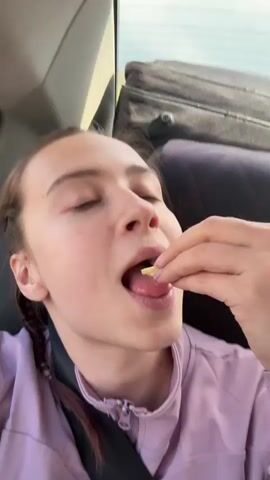 A young woman with a braid in a light purple zip-up jacket is seen inside a car, likely in the back seat, wearing a seatbelt. She is eating small, yellow pieces of food, which appear to be popcorn or corn snacks, placing them into her mouth. Her expression is neutral, and she is focused on eating. Behind her, a car window shows bright daylight, and a black bag is visible on the seat. The setting suggests she is traveling in a car. No text is visible in the video. The activity takes place in Горняк, Russia.