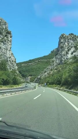 The video shows a first-person perspective from inside a vehicle driving on a highway through a mountainous landscape. The highway has multiple lanes and is bordered by guardrails. Steep, rocky cliffs covered in green vegetation rise on either side of the road. The sky is clear and blue with a few wispy pink clouds visible. Several other vehicles, including cars and possibly a truck, are visible on the highway ahead. Road signs are present along the side of the road. The sun is shining, indicating it is daytime.
