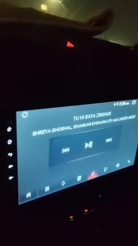 The video is filmed from inside a car at night, looking out through the windshield. The car's dashboard is visible at the bottom of the frame, including a screen displaying music information. The screen shows song title "TU HI BATA ZINDAGI" and artists "SHREYA GHOSHAL, SHANKAR EHSAAN LOY, and JAVED ALI". The car appears to be driving on a wet road during what seems to be a storm, with visible rain and wind affecting the trees and foliage outside. Streetlights and other lights are visible in the distance, reflecting on the wet surface. There are no people clearly visible in the video.
