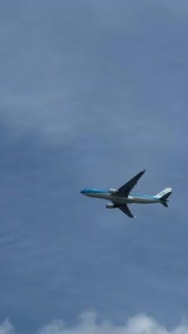 A KLM airplane flies across a clear blue sky. The airplane is viewed from the side, with its wings and engines visible. The KLM logo is visible on the tail of the plane. The sky is bright and appears to be daytime.