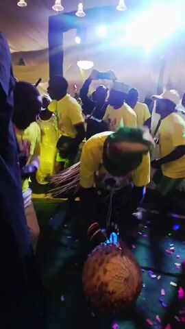 The scene captures a crowded indoor event where many individuals are participating in a lively celebration. Most of the attendees are wearing bright yellow T-shirts with a design on them. Among them, one person stands out due to their unique attire – a green head covering and a posture leaning forward, indicating engagement with the performance. The crowd is moving and gesturing, suggesting a joyful and energetic atmosphere. Colorful confetti litters the floor, adding to the celebratory mood. The lighting is bright, with spotlights overhead illuminating the event.