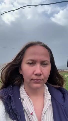 Here's a description of the video:
A woman stands outside in what appears to be light snow or sleet, filming herself. She has long brown hair, fair skin, and wears a light-colored, striped shirt under a dark blue vest with a patterned collar. Some snowflakes are visible on her face and hair.

Behind her is a green lawn, several parked cars, and buildings that look like houses or cabins. Distant mountains are visible in the background. The sky is overcast. The camera remains relatively still as she looks directly at the lens.