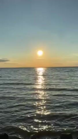 The video depicts a view of a body of water, likely a lake or sea, at sunset. The sun is a bright, golden orb positioned just above the horizon line, casting a shimmering reflection on the water's surface. Gentle ripples and small waves disturb the water, creating a textured, glittering effect. The sky is a gradient of light blue at the top, transitioning to a warmer orange near the horizon. A faint line of clouds is visible at the horizon. The foreground shows some darker shapes that might be rocks or some kind of barrier along the shore. The scene is serene and still.