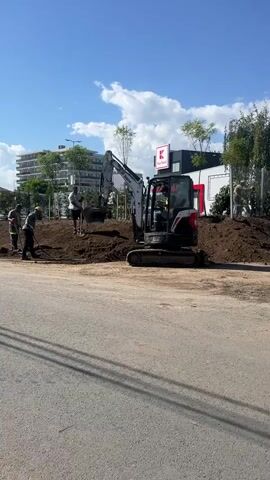 This is a short, vertical video clip showing an outdoor scene under a clear blue sky with scattered clouds. The foreground is dominated by a paved road with visible lane markings. Beyond the road, a construction site is in progress. A small, white Bobcat excavator is positioned on a mound of dirt, its bucket raised. A person, likely the operator, is visible inside the excavator's cabin, wearing a high-visibility vest and a hat.

To the left of the excavator, three construction workers are actively engaged in work. Two of them are using shovels to move or level the dirt. One worker is wearing a yellow hard hat and a light-colored shirt with a reflective vest. Another worker is wearing a dark shirt and dark pants. A third worker, further left, is partially obscured but appears to be interacting with the others or observing the work.

In the background, there are modern buildings, some with balconies, suggesting an urban or suburban environment. A prominent red and white "K" logo, likely from a retail store, is visible on a dark building. Trees and fencing are also present, indicating landscaping or property boundaries. The overall impression is a construction or landscaping project in progress on a sunny day. The video captures the movement and activity of the workers and the excavator.