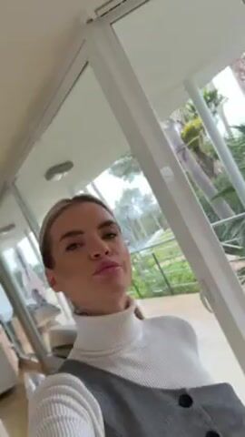 A woman with blonde hair in a ponytail, wearing a white turtleneck and a gray dress, is standing inside a room with large windows. The camera is at a low angle, looking up at her. The woman looks at the camera, turns her head slightly, and then looks back at the camera. Outside the windows, there are green plants and trees. There is a hint of a building in the background. The overall impression is that the woman is posing for the camera in a well-lit, modern space.