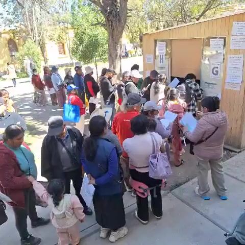 Certainly! Here's a concise description of the video:

The video shows a crowd of people standing in lines outdoors on a sunny day. They appear to be waiting for something near a small wooden building or booth marked with letters "H" and "G". Most individuals are holding papers.  Some are wearing masks.  Some are seated in blue plastic chairs. The surrounding environment includes trees and what looks like a multi-story building in the background. The camera moves slightly, showing the length of the lines. There appears to be a park-like area.