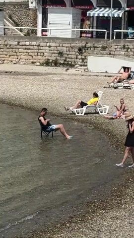 The video shows a beach on a sunny day. The beach is pebbly and crowded with people sunbathing on chairs and towels. A man sits on a folding chair in the water by the shore, looking at his phone. A woman in a black dress and straw hat walks by him on the beach. Behind them is a stone wall with buildings and shops above it. The camera zooms out to show more of the beach and the buildings in the background.