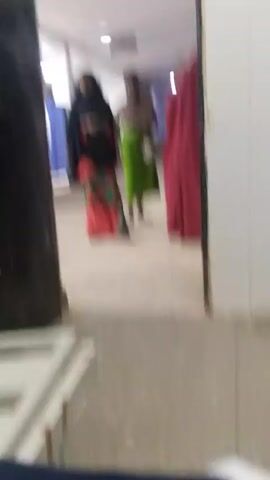 The video depicts a hallway scene with three individuals visible. On the left, a woman wearing a black hijab, a patterned red and green skirt, and a black top stands facing towards the right. In the center, another woman with her hair tied up, wearing a brown top and a long green skirt, faces forward. To the right, a third person, partially obscured by a doorway, is dressed in a red garment. The hallway has tiled floors and white walls, with a glimpse of blue curtains in the background. A white metal frame is visible in the foreground. The lighting appears to be fluorescent.