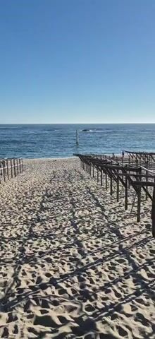 The video shows a beach with sand and the ocean in the background. There are wooden structures on the sand, which appear to be some type of seating or canopy frames, arranged in rows. The structures cast shadows on the sand. In the distance, in the water, there is a white pole with a red top, possibly a marker or a small lighthouse. The sky is clear and blue. There are no people visible in the immediate foreground, but there appear to be a few people standing in the distance near the wooden structures on the right side of the frame. The waves are gently rolling onto the shore.