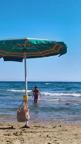 A person wearing a brown bikini walks into the ocean from a sandy beach. A colorful striped umbrella is in the foreground, with a brown bag hanging from its pole. Two people are in the water, appearing to be walking away from the shore. The sky is clear and blue, and the water is calm with small waves. The overall scene depicts a sunny day at the beach.