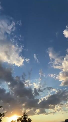 A time-lapse video shows clouds moving across a bright blue sky. The sun is visible on the left side of the frame, casting a warm glow. Trees are silhouetted against the horizon at the bottom of the image.