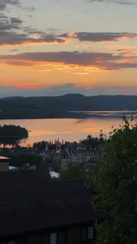 This is a vertical, eye-level, medium-range shot of a picturesque sunset over a lake in Mont-Tremblant, Canada. The sky is a canvas of warm, vibrant colors, with streaks of orange, pink, and gold painting the clouds. The setting sun casts a warm, reflective glow on the calm water of the lake, creating a mirror-like effect that doubles the beauty of the sky.

In the middle ground, a quaint village nestled by the lake comes into view. Its charming architecture, characterized by peaked roofs and intricate details, is bathed in the soft, warm light of the sunset. A prominent clock tower stands tall, adding to the village's idyllic charm. Surrounding the village are lush green trees and rolling hills, forming a natural backdrop that enhances the serene beauty of the scene. On the left side of the lake, a small, tree-covered island adds to the picturesque landscape.

In the foreground, the dark, textured roofs of buildings create a sense of depth and perspective, framing the view of the village and the lake. The leaves of a tree in the lower right corner add a touch of nature's vibrancy to the composition.

The overall mood of the image is one of tranquility and serenity. The warm colors of the sunset evoke a sense of peace and contentment, while the beautiful scenery inspires awe and wonder. The scene captures the magic of a summer evening in Mont-Tremblant, a perfect moment of natural beauty and charming village life. The image does not contain any visible text.