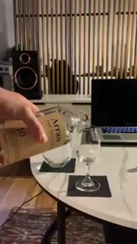 A person pours Arran 10 single malt scotch from a glass bottle into a stemmed glass on a white round table. A water glass with ice and a laptop are also on the table, which is placed in front of a decorative bamboo screen. A black speaker is visible in the background, along with a bit of an area rug and other furniture.