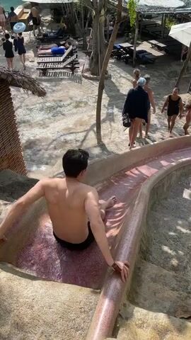The video shows a small outdoor water park area. A shirtless man is sitting on the edge of a pink water slide, ready to slide down. The slide curves into a green-tinted pool of water. Other people are standing around the pool and some are in the water. Some people are walking around the edge of the pool, and a small child is also visible.  White umbrellas and thatched-roof structures provide shade in the background, where there are lounge chairs. The area is surrounded by trees. The man slides down the slide and into the pool, creating a splash.