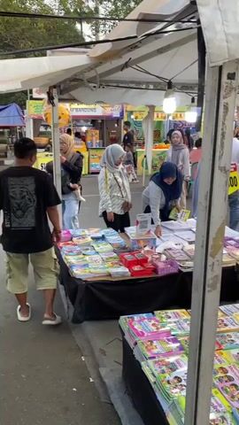 A street market stall displays stacks of children's books. A person wearing a black t-shirt and shorts is browsing the books on a table. Another person, wearing a light-colored hijab and a white patterned top, is looking at books. A third person, wearing a blue hijab and a grey top, is arranging books on a table. Further back, several other people are walking around and browsing other stalls. The stall is covered by a white canopy, and there are lights hanging from it. Price signs are visible, including one that reads "10.000 dpt 3" and another that reads "5.000". The ground is paved.