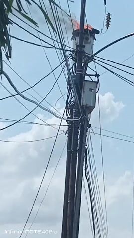 The video shows a power pole with an electrical transformer on fire. Flames are visible coming from the top of the transformer, and black smoke is rising. The pole is surrounded by a dense network of electrical wires. The sky is blue with some clouds in the background. The footage appears to have been taken from ground level, looking up at the pole. At the bottom of the image is the text, "Infinix NOTE 40 Pro".